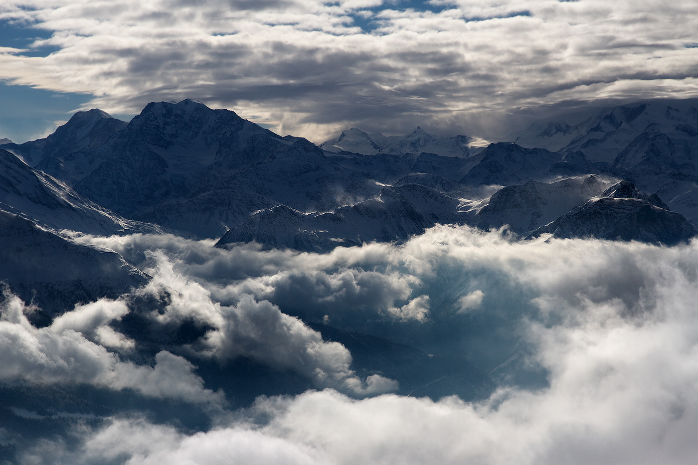 A sweeping mountain landscape rises above a sea of clouds, with jagged, snow-covered peaks stretching across the horizon. The light is soft and diffused, filtering through layered clouds that drift between the ridges, partially veiling the valleys below. The mountains appear dark and solid against the shifting البيضاء-white clouds, their contours highlighted by patches of snow and subtle glints of light.
The scene feels vast, powerful, and almost untouchable. There’s a sense of elevation and freedom—as if standing at the edge of the sky itself—where only birds like eagles might roam. Emotionally, it evokes awe and quiet reverence, mixed with a hint of solitude. The interplay of light and shadow creates a contemplative mood, suggesting both the grandeur of nature and the smallness of the observer within it.