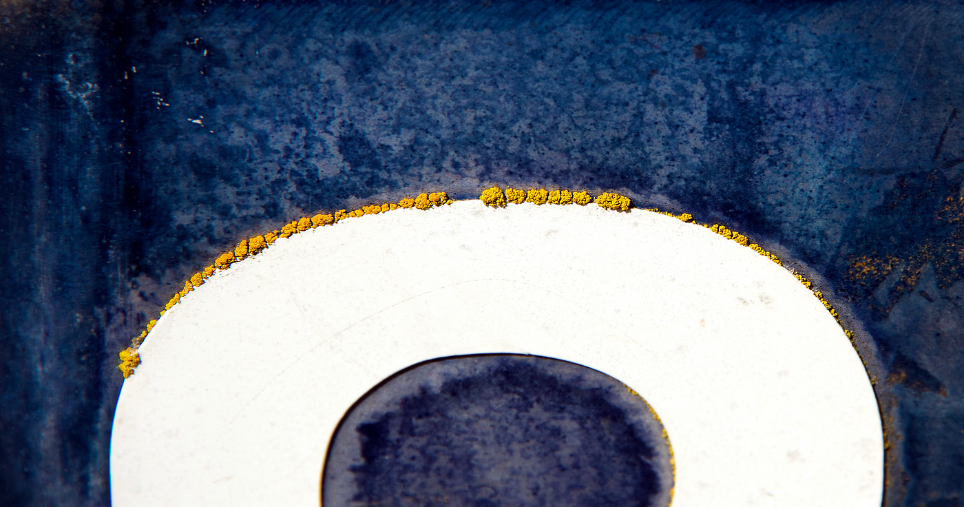 A close-up, abstract composition centers on a smooth, pale circular form set against a darker, textured blue-gray background. Along the upper edge of the circle, a thin line of small, vivid yellow clusters—possibly lichen or mineral deposits—traces the curve like a delicate border. The lower portion of the circle contains a darker, oval shadow or recess, adding depth and contrast. The surrounding surface appears worn and slightly mottled, with subtle scratches and variations in tone.
The image feels minimal yet striking. The strong contrast between the cool blues and the bright yellow creates a quiet tension, while the clean curve of the circle introduces a sense of order within an otherwise weathered, organic setting. Emotionally, it evokes curiosity and contemplation—like a small, overlooked detail transformed into something almost cosmic or symbolic. There’s a calm stillness, paired with a sense of intrigue, as if hinting at hidden processes or slow, natural change.