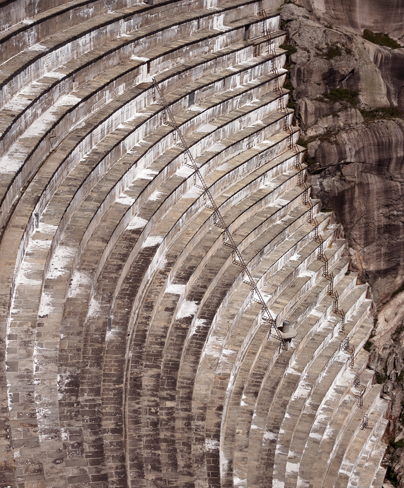 A massive curved concrete structure fills the frame—likely part of a dam—its surface marked by repeating horizontal tiers that arc gracefully across the image. Each level is weathered with pale streaks and mineral stains, giving the structure a textured, aged appearance. Narrow staircases zigzag vertically along the curve, connecting the tiers with a precise, almost geometric rhythm. On the right edge, rugged natural rock contrasts with the engineered form, emphasizing the boundary between nature and human construction.

The image feels monumental and slightly overwhelming. The repeating lines and sheer scale create a sense of order and control, yet also hint at the effort and endurance behind such a structure. Emotionally, it evokes awe mixed with a quiet tension—the kind that comes from standing before something vast and immovable. There’s also a contemplative quality, as if the steps suggest a long, deliberate journey, emphasizing persistence, time, and human ambition.