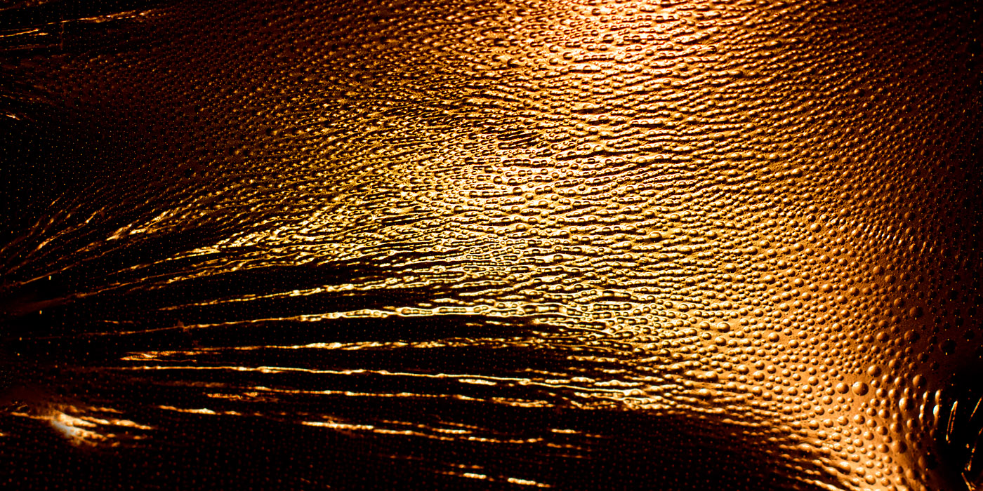 A richly textured surface fills the frame, glowing in deep amber and golden tones. Countless tiny droplets bead across it, catching the light and forming intricate patterns—some clustered densely, others stretched into streaks as if pulled by motion. The surface beneath appears smooth yet subtly contoured, with flowing lines that guide the eye diagonally across the image, creating a sense of movement within stillness.

The scene feels warm, almost tactile, and slightly abstract—like a close-up of skin, metal, or glass transformed into something unfamiliar. The golden light gives it a sensual, almost primal quality, while the repetitive droplets add a rhythmic calm. Emotionally, it evokes a blend of intimacy and intensity—both soothing and charged—suggesting heat, closeness, and a quiet, underlying energy.