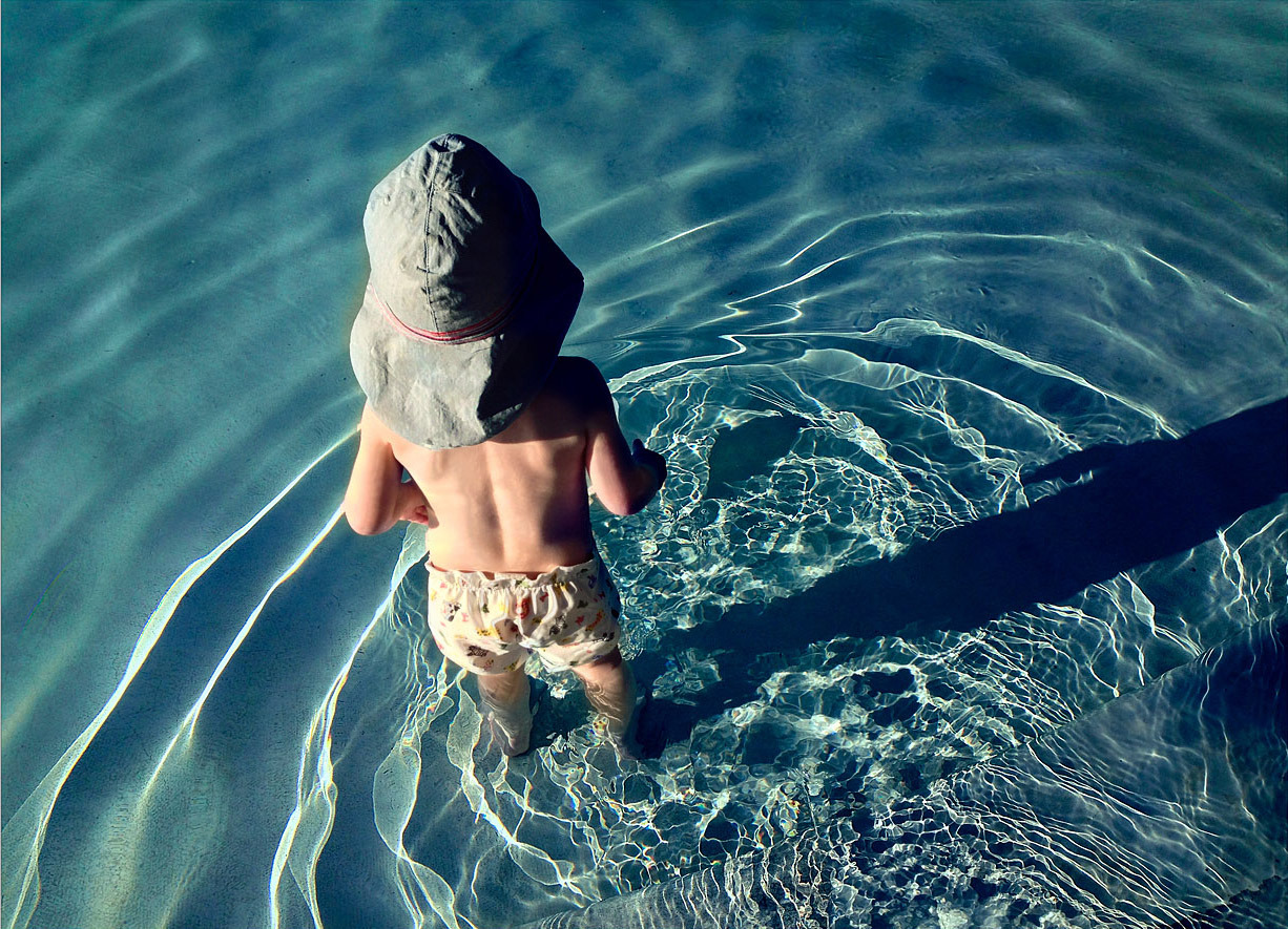 A small child stands ankle-deep in clear, gently rippling water, seen from above. They wear a light-colored sunhat that shades their head and a pair of patterned shorts, their bare back catching the sunlight. The water is a luminous blue-green, with bright, wavy reflections dancing across the surface and around the child’s legs. A long, dark shadow stretches beside them, distorted by the moving water.

The scene feels warm, simple, and quietly joyful. There’s a sense of innocence and curiosity in the child’s stance, as if they’re absorbed in the small wonder of the water around them. The shimmering light and soft movement create a peaceful, almost dreamlike atmosphere. Emotionally, it evokes nostalgia and tenderness—a fleeting moment of childhood exploration, full of calm, wonder, and gentle freedom.