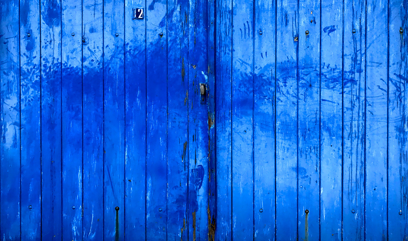 A large wooden door painted in vivid blue fills the frame, its vertical planks worn and weathered over time. The paint is uneven, with darker streaks, smudges, and faded patches revealing age and use. Near the top, a small number “2” is fixed slightly off-center, and a simple metal handle sits at about mid-height. Subtle marks, scratches, and drips run down the surface, adding layers of texture and history.
The image feels quiet but full of character. The bold blue color gives it a strong, almost confident presence, while the wear and imperfections suggest time, memory, and stories untold. Emotionally, it evokes curiosity and nostalgia—like a door that has witnessed countless moments, inviting you to imagine what lies behind it.