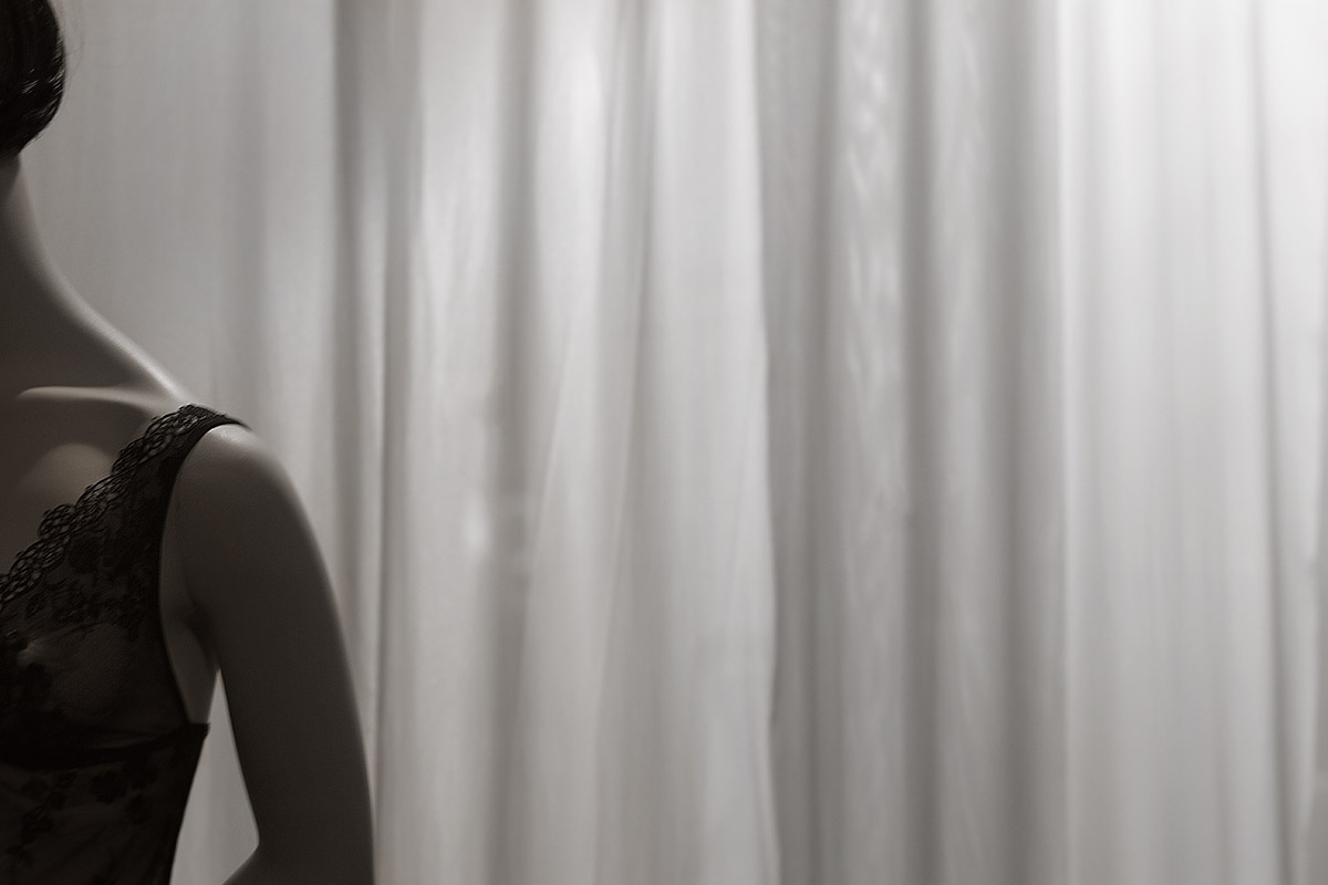 A softly lit, minimalist scene shows the partial figure of a person standing at the edge of the frame, their body turned slightly away so that only the shoulder, neck, and a hint of the face are visible. They wear a dark, delicate lace garment that contrasts with the pale, smooth tones of their skin. Behind them, sheer white curtains hang in gentle vertical folds, diffusing the light and creating a calm, muted backdrop.

The composition feels intimate and restrained, with much of the image left open and quiet. The soft lighting and limited view of the figure create a sense of privacy and subtle mystery, as if the moment is personal and unobserved. Emotionally, it evokes calmness and introspection, with a delicate tension between vulnerability and elegance—like a quiet pause before or after something meaningful.