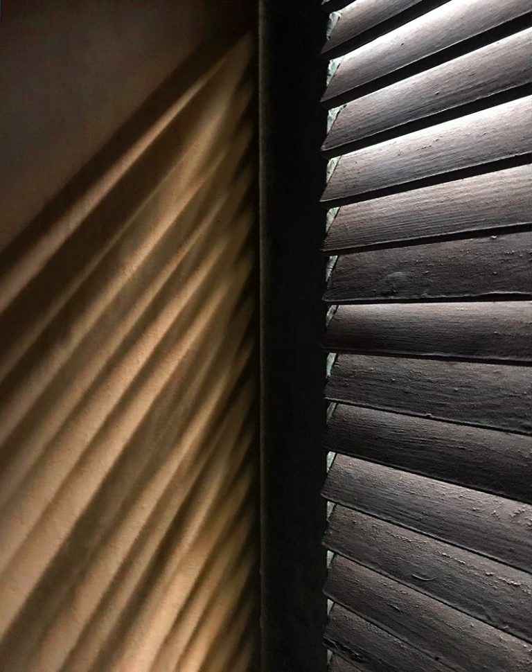 Light slips through narrow blinds, casting quiet stripes across the wall like passing time made visible. Shadows and brightness weave together, creating a hushed, intimate rhythm—half hidden, half revealed—where stillness lingers and the ordinary becomes contemplative, holding a fragile balance between secrecy, warmth, and the soft whisper of day.