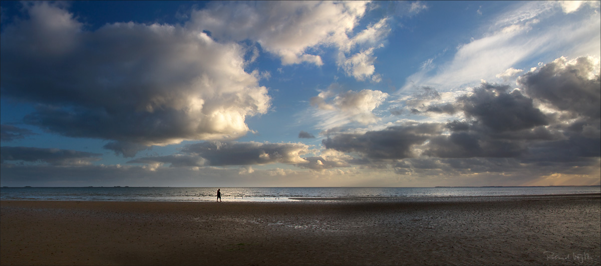 Dramatic rays of light pierce heavy clouds above a lush green Swiss valley meadow. Wisps of mist drift through a dark conifer forest on the horizon. A solitary farmhouse sits to the right. The mood is charged – suspended between storm and stillness, between shadow and sudden illumination.