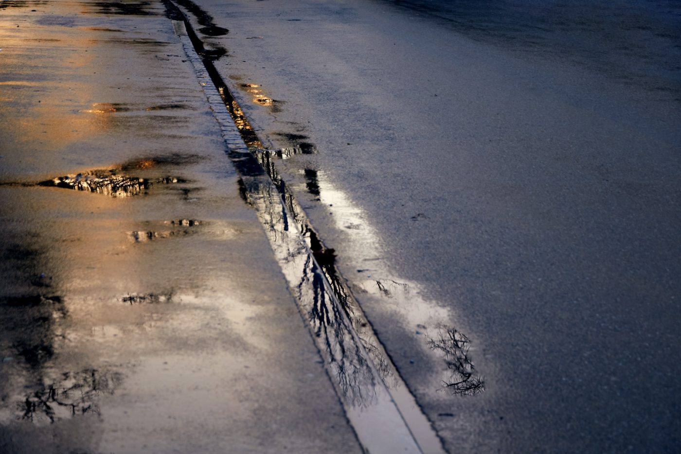 (english alt-text follows)

Eine nasse Asphaltstraße im schwindenden Licht, durchzogen von einer schmalen Fuge, in der sich Regenwasser sammelt. Kleine Pfützen spiegeln den Himmel und die kahlen Äste von Bäumen, als läge eine stille, zerbrechliche Welt unter der Oberfläche. Warmes Licht glimmt in einzelnen Reflexionen, während der Rest in kühlem Blau und Grau verharrt. Die Szene wirkt ruhig und melancholisch — ein Moment zwischen Vergehen und Verweilen, in dem sich Tiefe im Unscheinbaren offenbart.

EN :
A wet asphalt road in fading light, marked by a narrow seam where rainwater gathers. Small puddles reflect the sky and the bare branches of trees, as if a quiet, fragile world lies just beneath the surface. Patches of warm light glow within the reflections, while the rest lingers in cool blues and greys. The scene feels still and melancholic — a moment between passing and staying, where depth reveals itself in the ordinary.