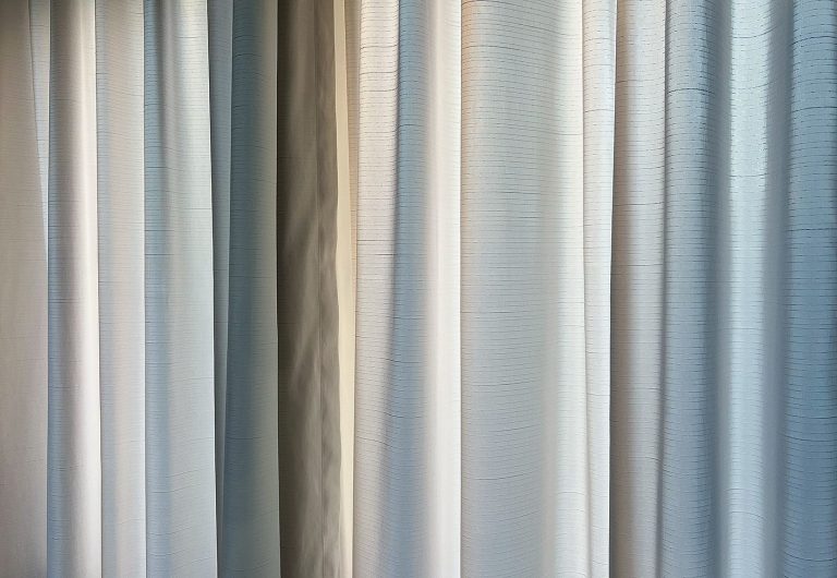 A sheer white curtain fills the frame, illuminated by soft morning light filtering through the fabric. Delicate folds create subtle patterns of shadow and texture, revealing gentle transitions between white, gray, and faint hints of blue. The image is quiet and minimalist. What first appears to be an ordinary curtain gradually unfolds into a rich landscape of light, depth, and detail, inviting a moment of still attention.