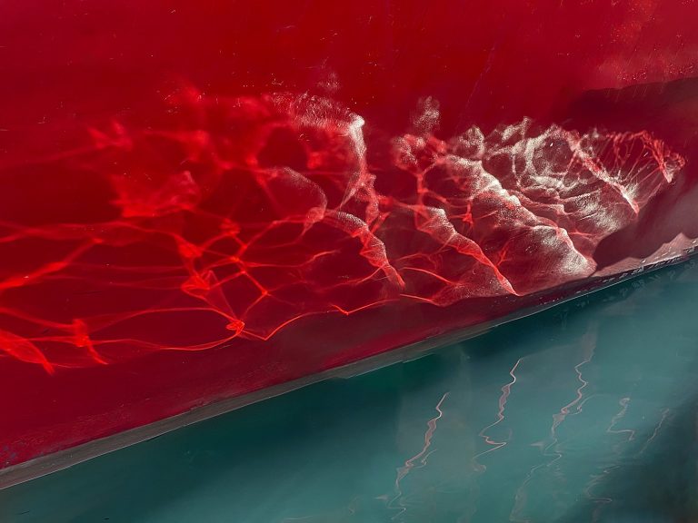 Deutsch Ein roter Schiffsrumpf trifft auf dunkles, türkisfarbenes Wasser. Sonnenlicht bricht sich an der Wasseroberfläche und wirft helle, verzweigte Lichtbänder an die Bordwand. Wo sich die Spuren berühren, entstehen weisse Verdichtungen. Unterhalb der Wasserlinie spiegeln sich schwache rote Schlieren im Grün der Tiefe. Die Bühne eines Tanzes, der noch leuchtet — und bereits vorbei ist. English A red ship's hull meets dark turquoise water. Sunlight refracts at the surface and throws bright, branching bands of light against the hull. Where the traces touch, white intensities form. Below the waterline, faint red streaks reflect in the green of the depths. The stage of a dance still glowing — and already gone.