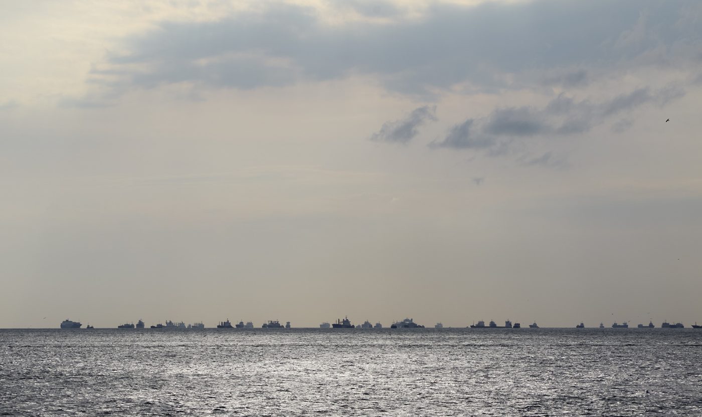 Zahlreiche Frachtschiffe liegen weit verstreut auf einer ruhigen, graublauen Meeresfläche. Im Dunst verschwimmen ihre dunklen Silhouetten mit dem Horizont; im Hintergrund sind schemenhaft Küste, Hafenanlagen und Stadt zu erkennen. Die Szene wirkt still und erwartungsvoll, als warteten die Schiffe vor einem unsichtbaren Hafen auf Einfahrt.
Numerous cargo ships are scattered across a calm, blue-grey sea. Their dark silhouettes fade into the misty horizon, while a distant coastline, port facilities and city skyline are barely visible in the background. The scene feels quiet and expectant, as though the ships were waiting outside an unseen harbour for permission to enter.