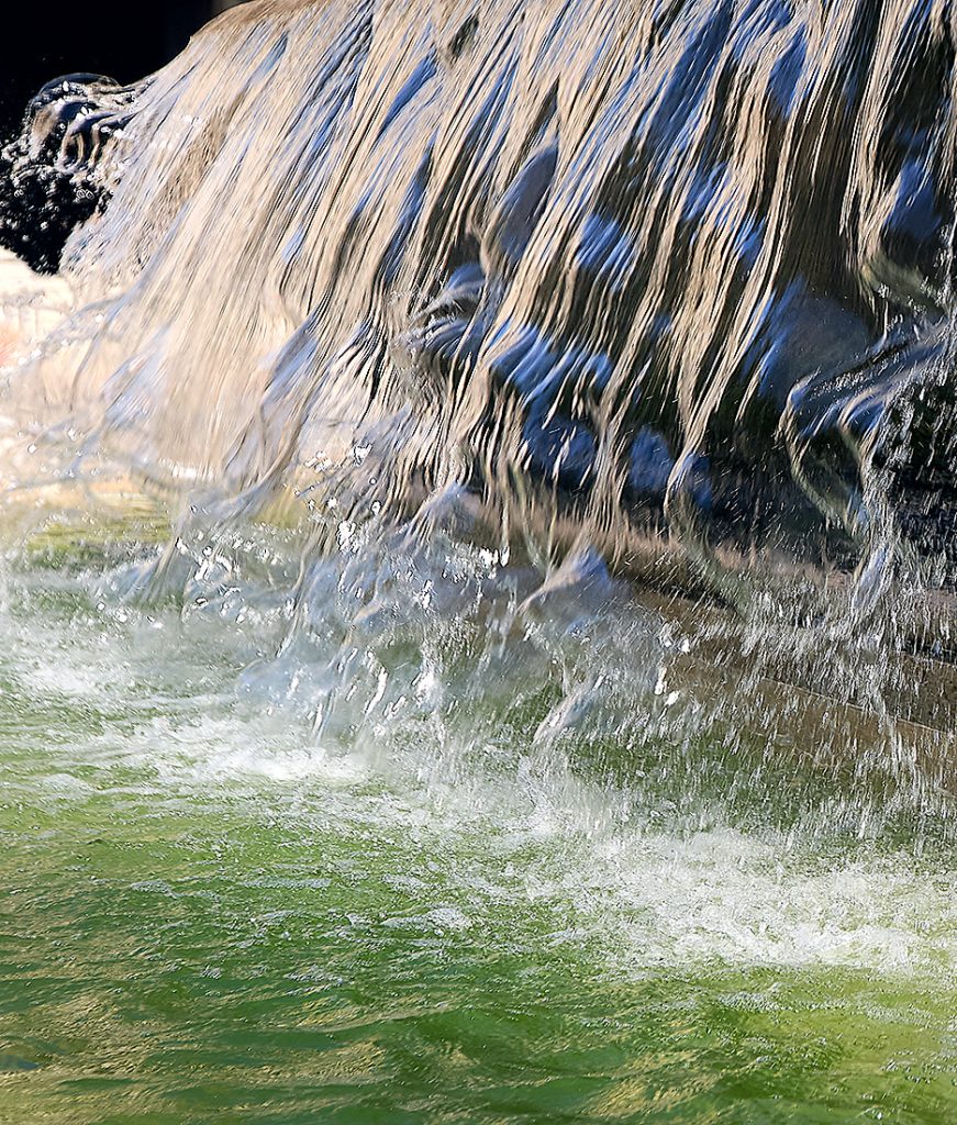 Wasser fällt in schmalen, lebendig gewundenen Strähnen über dunklen Stein. Der Brunnen wirkt wie das fliessende Haar einer Najade – kühl, zeitlos und voller Geschichten.